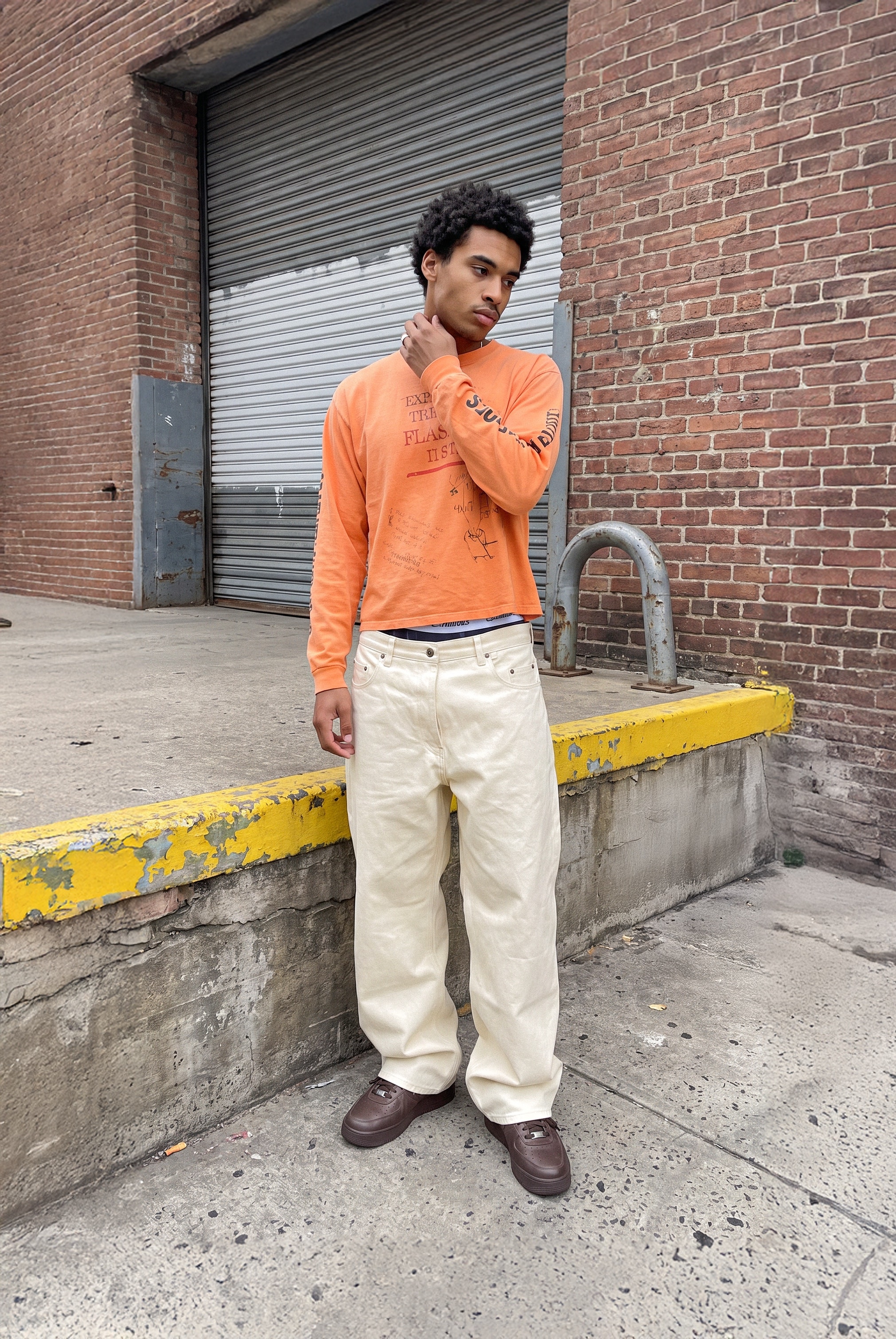 Model in an orange shirt and cream trousers by a loading dock